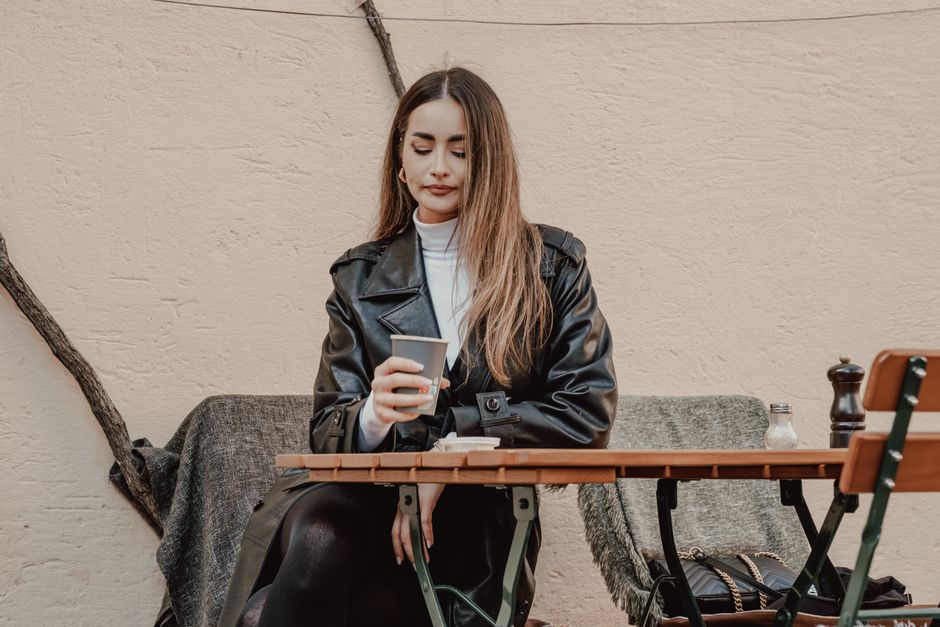 Stylish woman in leather coat sipping coffee at an outdoor café table.
