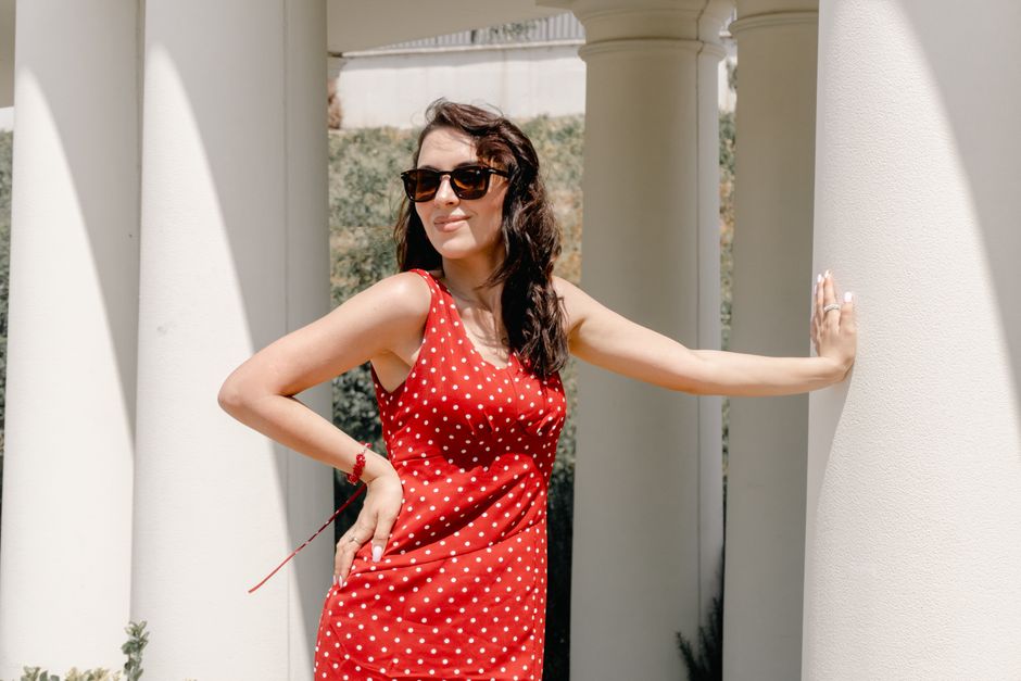 Stylish woman in a red polka dot dress poses confidently by columns under the sun.