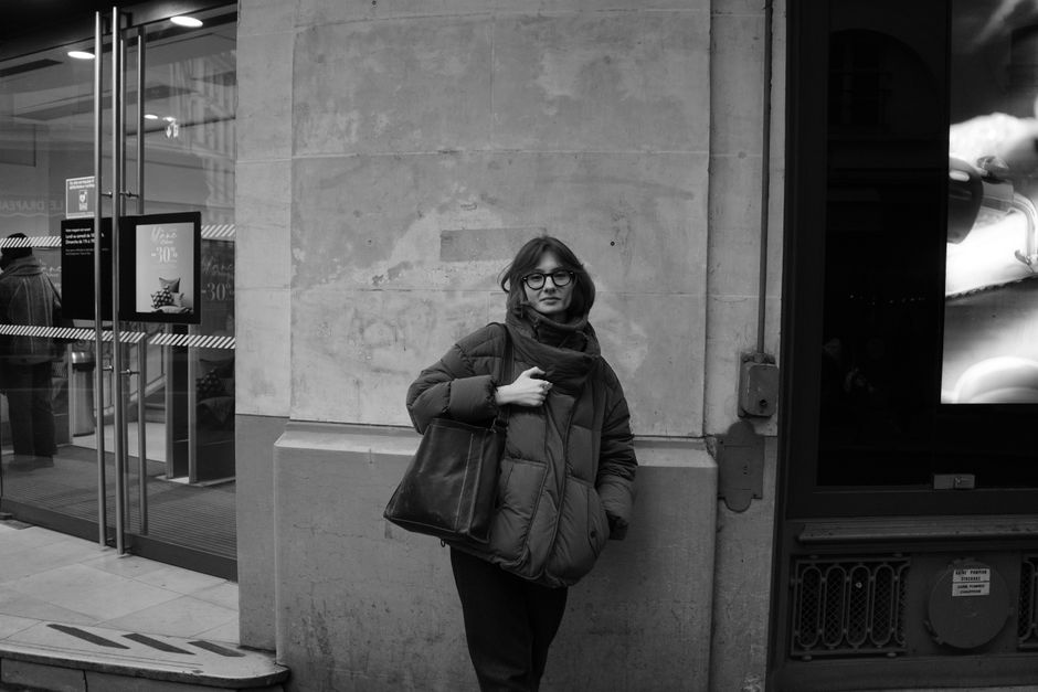 Black and white photo of a woman wearing a winter coat, standing by a storefront in a city.