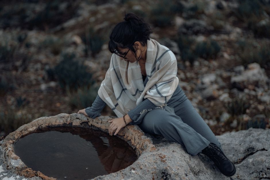 A woman in a scarf sits on rocks, gazing into a small stone pond.