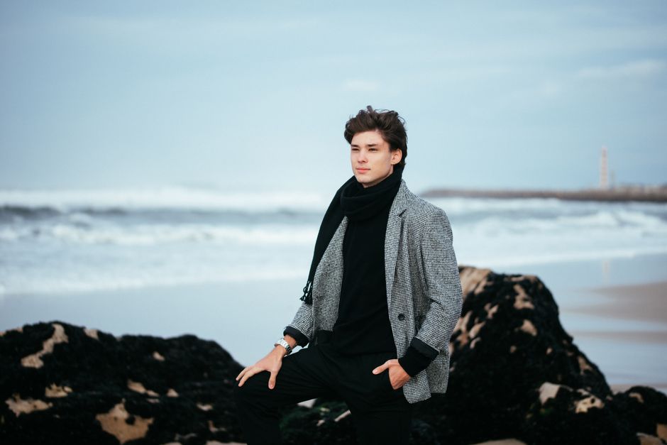 Stylish man in winter clothing posing on a beach with ocean waves in the background.