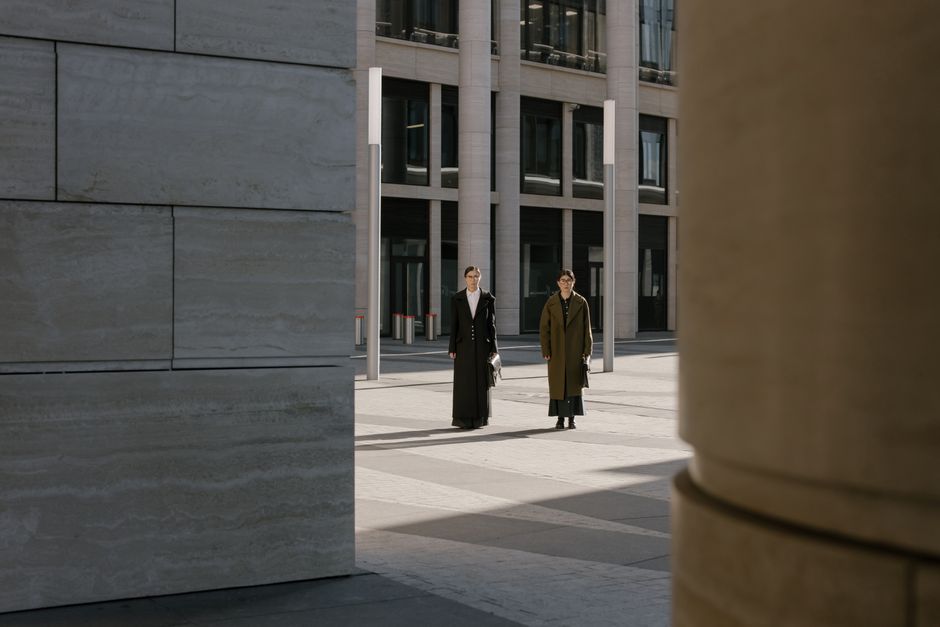 Two women in stylish overcoats walking in a modern urban architectural area during the day.