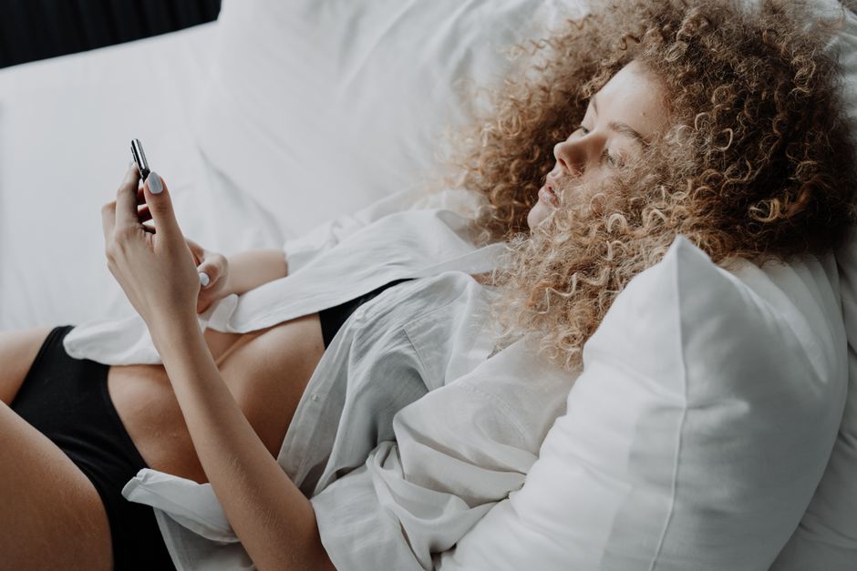 A woman with curly hair lies on a bed using her phone, wearing a white shirt.