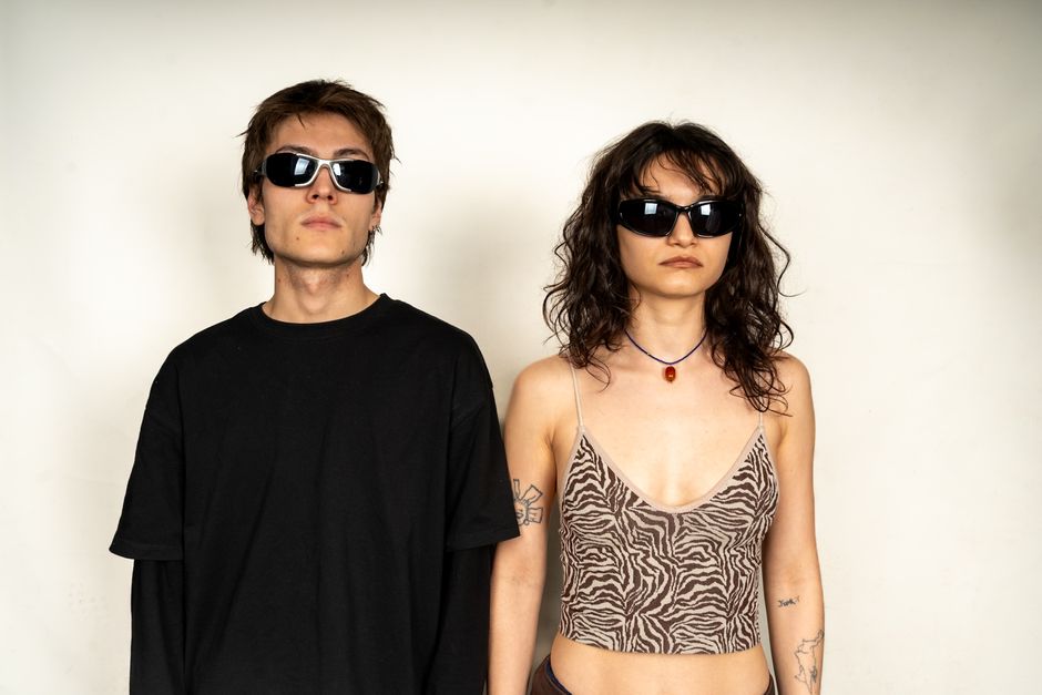 A fashionable portrait of two young adults wearing stylish sunglasses and modern attire against a plain background.