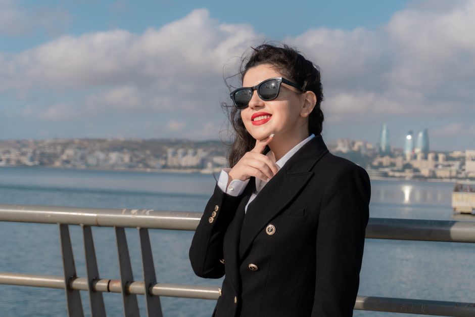 A young woman in stylish black attire smiles by the Baku seaside with a city skyline view.