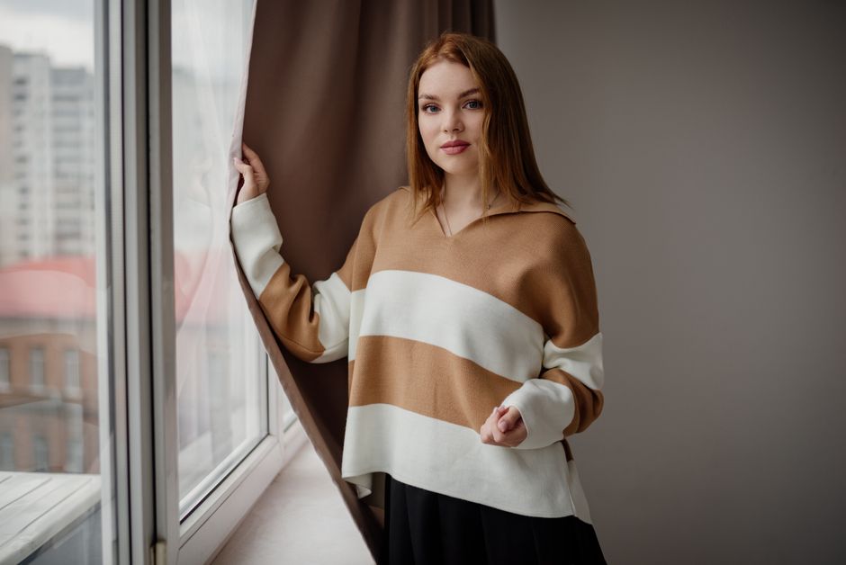 A young woman with red hair poses by a window in an urban apartment. Natural light enhances the cozy ambiance.