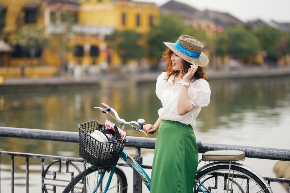 Woman posing with bicycle by a riverside in a quaint town, wearing a straw hat and green skirt.