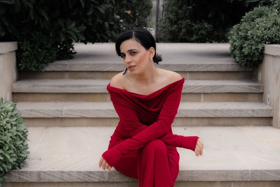 A stylish woman in a vibrant red dress sitting on stone steps surrounded by greenery.