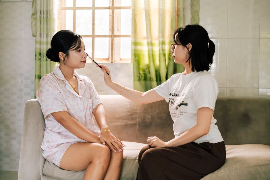 Two young women, one applying makeup on the other, seated on a sofa in a well-lit room.