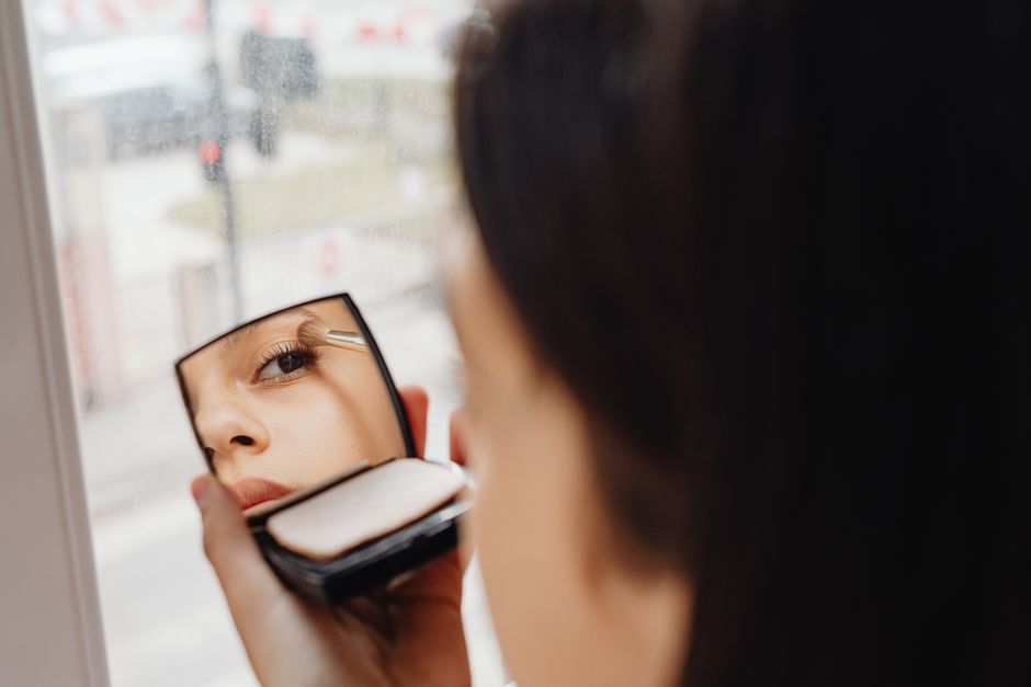 A woman applies eye makeup using a compact mirror, showcasing a close-up reflection effect indoors.